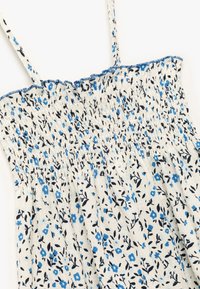 Vestido floral con fruncido que presenta pequeñas flores azules y hojas negras sobre un fondo crema claro, con tirantes delgados ajustables y bordes con volantes.