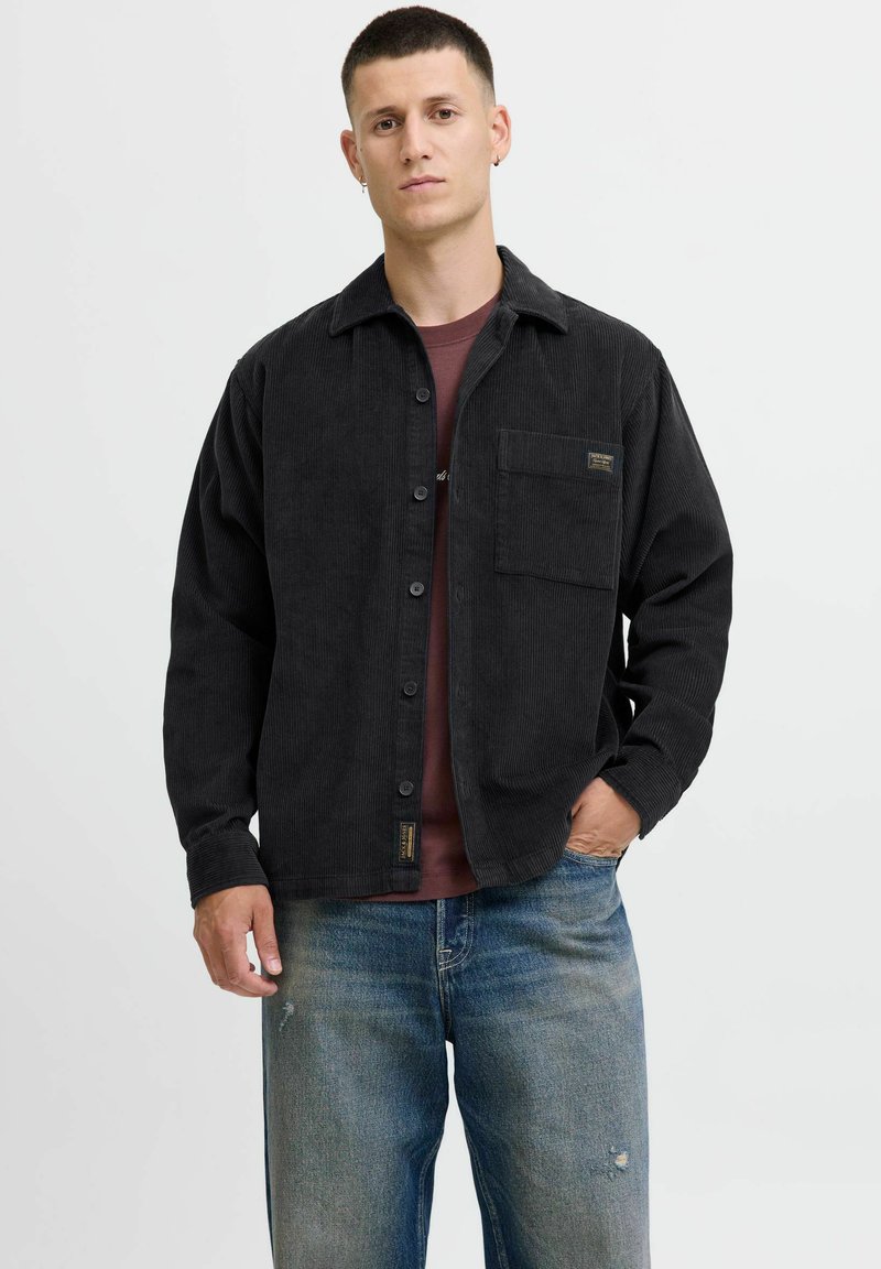Chemise en velours côtelé noire à boutons avec une poche poitrine, portée par-dessus une chemise bordeaux, associée à un jean déchiré bleu clair. Design simple.
