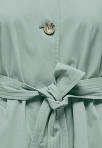 Tissu vert clair enveloppant avec une ceinture nouée, présentant un bouton rond à deux trous en haut. Texture lisse et design structuré.