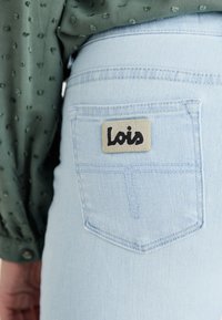 Svijetloplave traperice s stražnjim džepom na kojem se nalazi bež oznaka s natpisom "Lois" u crnoj boji, postavljene uz teksturiranu zelenu bluzu.