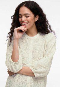 Light cream blouse with textured wave pattern, round neckline, and button closure. Puff sleeves with elastic cuffs. Lightweight fabric.