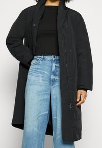 Chaqueta larga negra con un acabado texturizado, cierre de un solo snap y cremallera, llevada sobre un top negro de canalé y jeans de un azul claro.