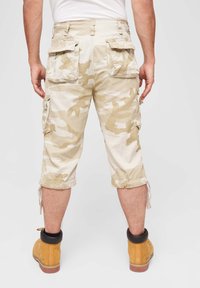 Knee-length cargo šortky v béžovom maskáčovom vzore, vyrobené z bavlny, s bočnými vreckami a nastaviteľnými šnúrkami na leme. Nosené s žltými čižmami.