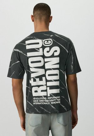 Muž nosiaci čierne tričko s bielym tie-dye vzorom, na ktorom je veľký biely vertikálny nápis „REVOLUTIONS“ a menší biely text v spodnej časti, skombinované s svetlomodrými džínsami.