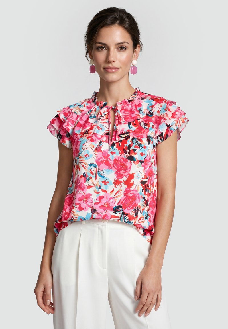 Femme portant un chemisier à manches courtes volantées avec un imprimé floral rose, rouge et bleu, un pantalon blanc et des boucles d’oreilles pendantes violettes et blanches.