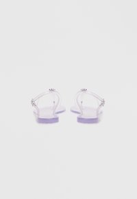 Gennemsigtige sandaler har en lilla gummisål, klare stropper og sølvfarvede metaldetaljer. Designet er minimalistisk og moderne.