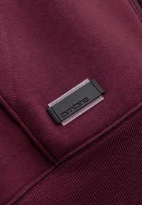 Primo piano di un tessuto rosso scuro con un'etichetta rettangolare in plastica nera e trasparente etichettata "ombre" cucita sul materiale.