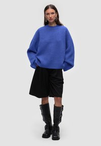 Blauer Wollpullover mit übergroßen Ärmeln, kombiniert mit schwarzen Shorts und knielangen schwarzen Stiefeln mit Schnallen. Schlichter Hintergrund.