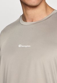 Ανοιχτό γκρι μπλουζάκι με στρογγυλή λαιμ όκοψη και κοντά μανίκια. Λευκό λογότυπο "Champion" τυπωμένο μπροστά, με ομαλή υφή.