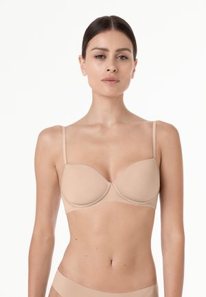 Donna con capelli scuri che indossa un reggiseno color nudo e biancheria intima abbinata su sfondo bianco uniforme, rivolta verso l'obiettivo con espressione neutra.