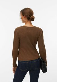 Femme aux cheveux bruns attachés en chignon, vêtue d'un haut marron à manches longues et d'un jean bleu foncé, tenant une pochette noire, dos à la caméra.