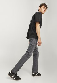 Svart T-skjorte med grafisk design, grå jeans og svarte joggesko med hvite detaljer; stående posisjon med en lett kroppsvending.