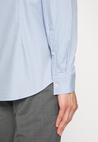 Lichtblauw overhemd met lange mouwen, knoopmanchetten en een gladde textuur. Grijze broek gedeeltelijk zichtbaar aan de onderkant, met een getailleerde pasvorm.