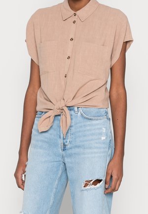 Camisa marrón claro de manga corta con botones, atada a la cintura, combinada con vaqueros desgarrados azul claro en una persona con tono de piel medio.