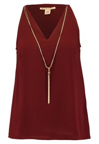 Burgundowa bluzka bez rękawów z dekoltem w kształcie V. Posiada złoty naszyjnik z długim frędzlem. Miękki, teksturowany materiał.