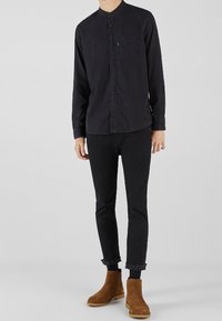 Camicia nera con bottoni e collo alla coreana, abbinata a pantaloni neri slim e stivaletti marroni, con taschino sul petto.