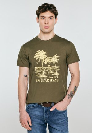 Młody mężczyzna z ciemnymi kręconymi włosami, ubrany w oliwkowo-zieloną koszulkę z nadrukiem palm, dżinsy i widoczne tatuaże na ramionach.