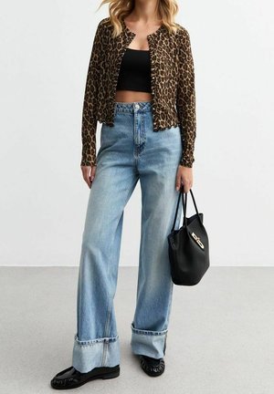 Frau trägt leopardgemusterten, geknöpften Cardigan, schwarzes Crop-Top, hoch taillierte, weit geschnittene Jeans, schwarze Loafers, hält schwarze Handtasche.