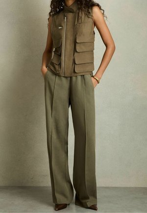 Femme portant un gilet sans manches vert olive avec de grandes poches, un pantalon large olive, des chaussures marron à bout pointu, et un bracelet en or, debout les mains dans les poches.