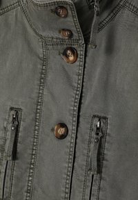 Chaqueta verde oliva con un alto cuello, cuatro bolsillos frontales, grandes botones marrones y detalles con cremallera. Tejido texturizado con costuras visibles.