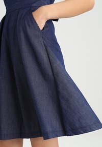 Džínové šaty s vypasovaným pasem a rozšířenou sukní. Mají boční kapsy, tmavě námořnickou barvu a hladkou texturu.