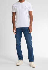 Camiseta blanca de algodón con mangas cortas, con un pequeño logo negro en el pecho. Combinada con pantalones vaqueros azules y zapatillas blancas.