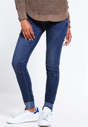 Personne portant un jean skinny bleu foncé avec des coutures diagonales, des revers roulés, des baskets blanches avec des accents bleus et un pull en tricot marron.