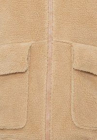 Casaco de fleece bege com fecho frontal, duas bolsos de pala e uma superfície texturizada. O design enfatiza o calor e o conforto.