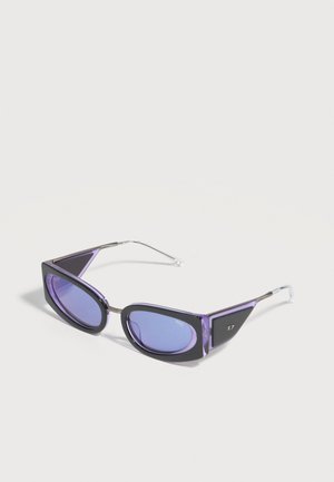 Οβάλ γυαλιά ηλίου με μοβ απόχρωση, με παχιά μαύρα και μοβ γωνιώδη πλαίσια και λεπτούς ασημένιους μεταλλικούς βραχίονες σε λευκό φόντο.