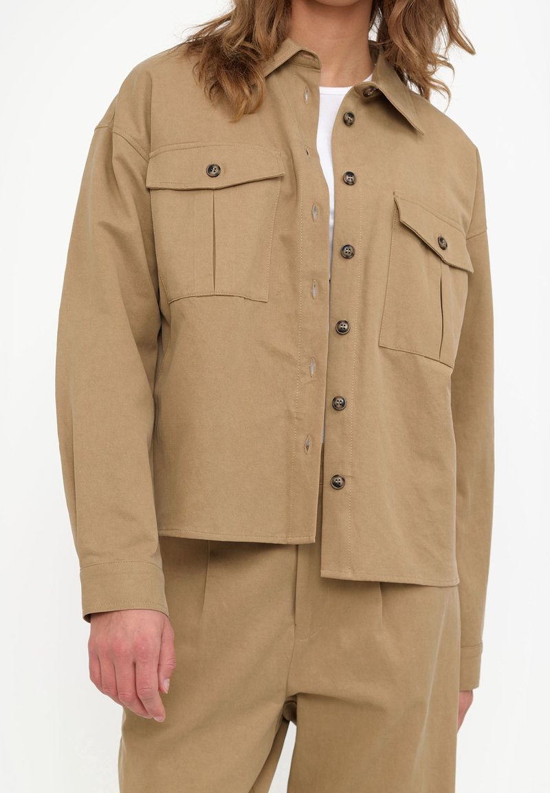 Femme portant une veste beige boutonnée avec des poches poitrine et un pantalon beige assorti sur fond blanc.