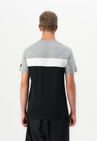 Mand, der er vendt væk, iført en kortærmet grå, hvid og sort farveblok t-shirt med sorte bukser foran en ensfarvet hvid baggrund.