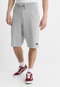 Mężczyzna w jasnoszarych szortach dresowych do kolan z troczkiem i małym prostokątnym logo na lewej nodze, w połączeniu z czerwonymi trampkami i białymi skarpetkami.