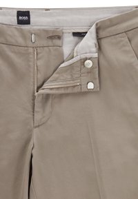 Pantalon en coton beige avec bouton et fermeture éclair, présentant une texture lisse et une légère brillance. Comprend une étiquette indiquant la marque.
