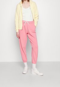 Světlomodrá mikina na zip, bílý top bez rukávů, růžové tepláky s důvlekem v pase a bílé tenisky, na jednobarevném pozadí.