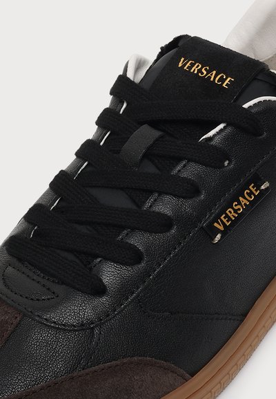 Baskets noires Versace avec lacets noirs, empeigne texturée, accents en daim marron et détails du logo dorés sur la languette et le côté.