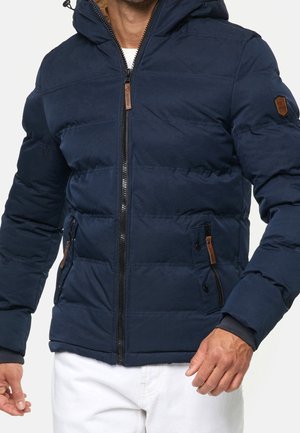 Marineblauwe pufferjack met gewatteerd ontwerp, capuchon, zwarte rits en tan leren accenten op de zakken. Heeft elastische manchetten en zijzakken.