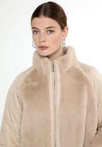 Giacca beige con zip, colletto in pelliccia sintetica morbida e pannello frontale; texture trapuntata sulle maniche; dettaglio della zip metallica.