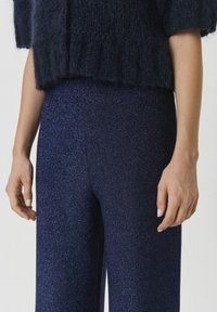 Marineblaue, glitzernde Hose mit weitem Bein, die eine glatte, glänzende Textur aufweist. Eine Hand ruht sanft auf dem Stoff und zeigt die Details.