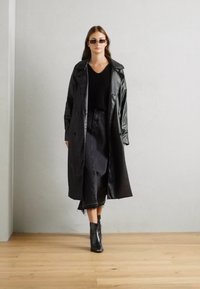 Manteau trench noir brillant sur un pull noir côtelé, associé à une jupe en jean et des bottines. Modèle présenté sur un fond neutre.