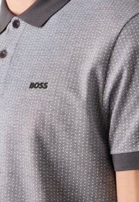 Πόλο μπλουζάκι σε γκρι ύφασμα με διακριτό μοτίβο κουκίδων. Διαθέτει σκούρο γιακά και τρία κουμπιά, με ένα μικρό μαύρο λογότυπο "BOSS" στο στήθος.