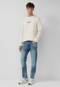 Camicia beige a maniche lunghe con logo di testo verde, jeans in denim blu e sneakers grigie con accenti bianchi, in piedi contro uno sfondo neutro.