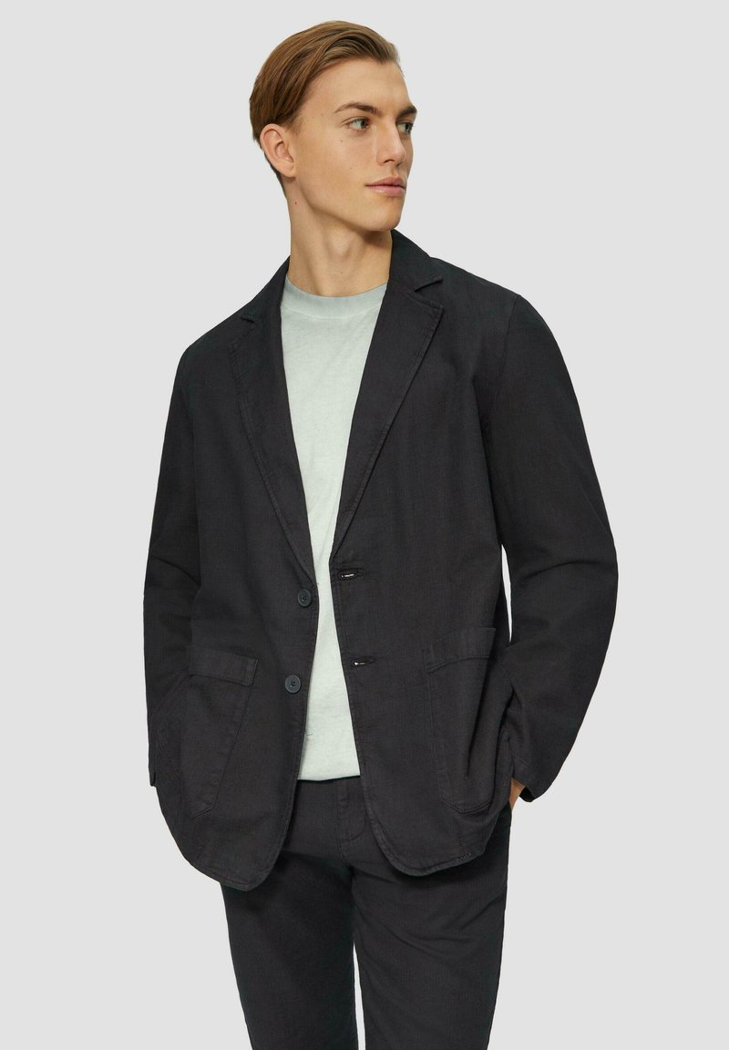 Schwarzer Leinenblazer mit notched Revers, zwei Fronttaschen und einer einreihigen Knopfschließung, getragen über einem hellgrauen T-Shirt.