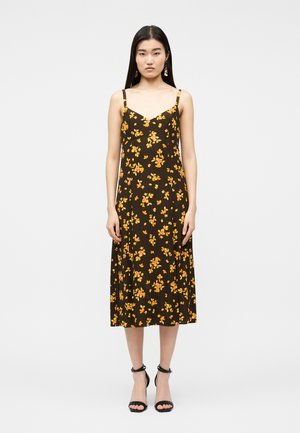 Femme aux longs cheveux noirs portant une robe midi marron à imprimé floral jaune et rose, avec des sandales noires à bride cheville, debout sur un fond blanc.