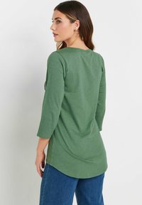 Top a maniche lunghe di colore verde oliva con scollatura rotonda, texture morbida e orlo leggermente asimmetrico, abbinato a pantaloni in denim azzurro chiaro.