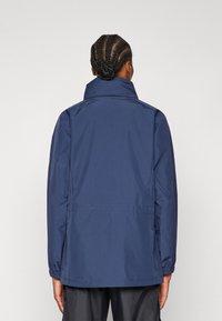 Navy blue waterproof jacket features a high collar, elastic cuffs, and a straight cut. The fabric has a smooth texture with minimal stitching.