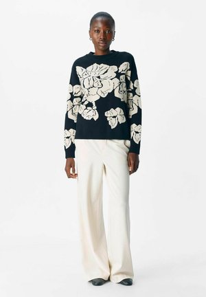 Zwarte trui met groot wit bloemmotief; gebreide textuur. Gepaard met crèmekleurige wijde broek en zwarte schoenen. Model poseert tegen een witte achtergrond.