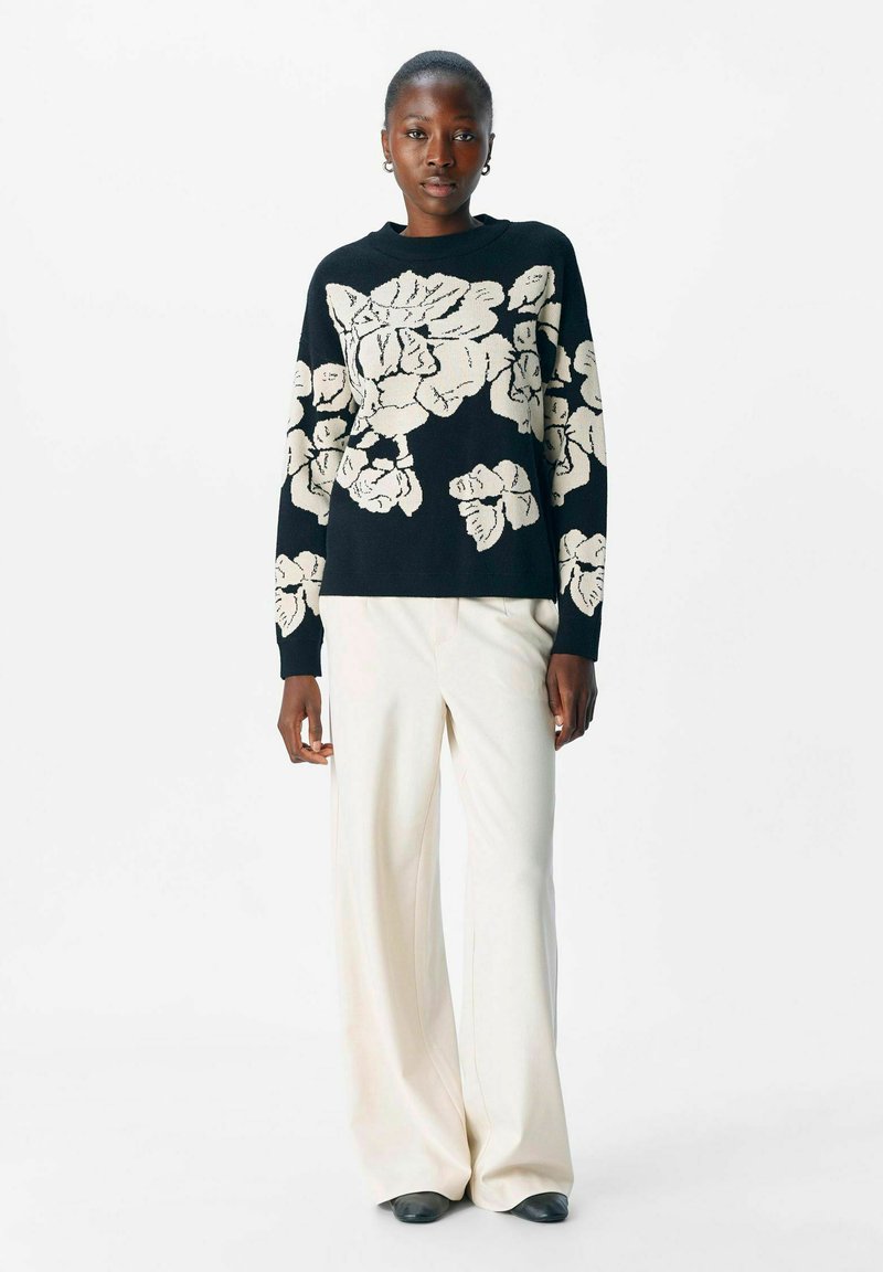 Suéter negro con un gran patrón floral blanco; textura de punto. Combinado con pantalones de pierna ancha crema y zapatos negros. Modelo posado contra un fondo blanco.