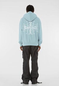 Svetlo modrá mikina s kapucňou a veľkým bielym grafickým dizajnom na chrbte, ktorý zobrazuje text "Hugo Mignonzales." Nosená s tmavými cargo nohavicami.