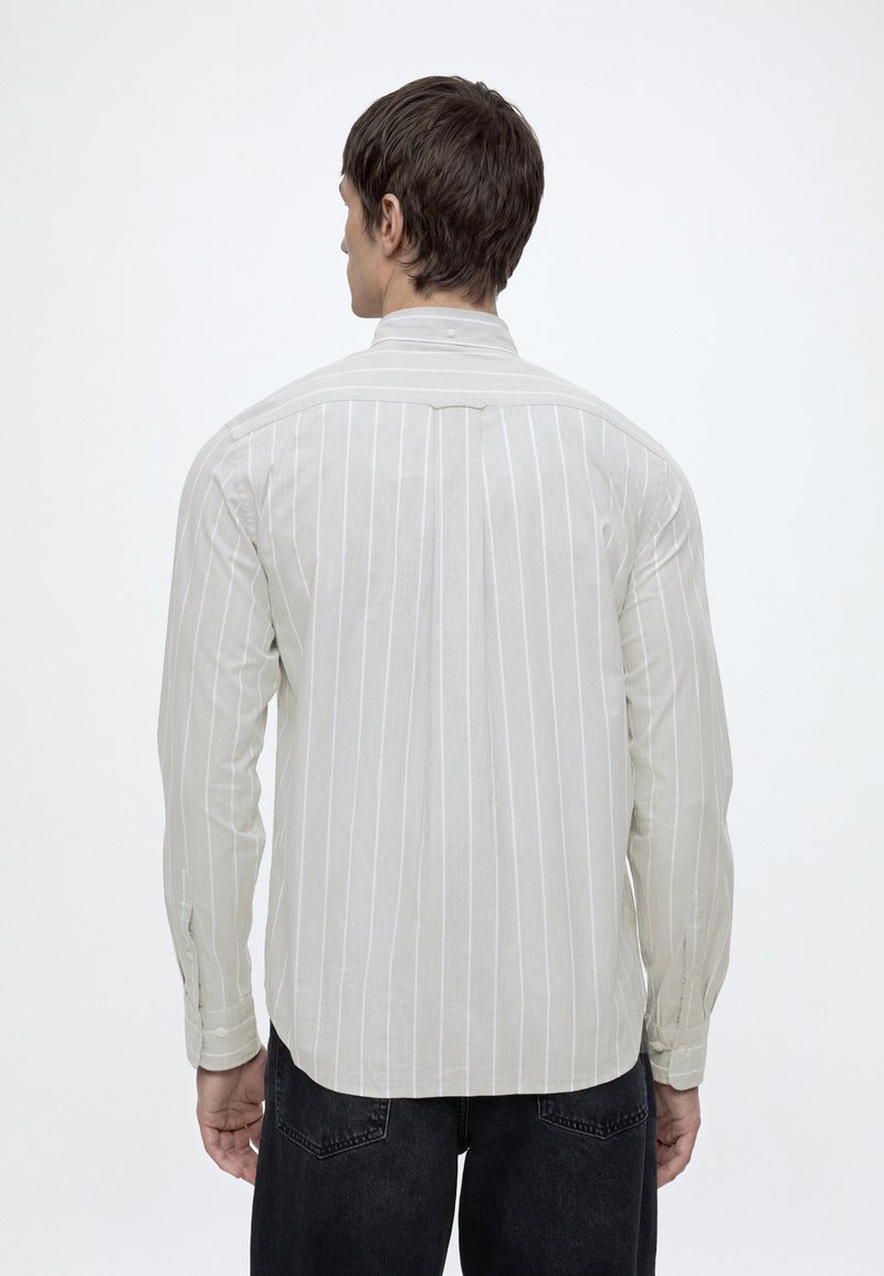 Mand iført lysegrå langærmet skjorte med hvide lodrette striber, stående med ryggen til, iført sorte jeans mod hvid baggrund.