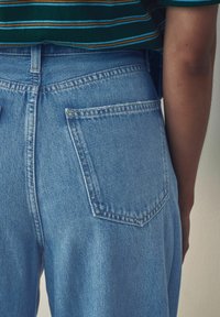 Jeans de denim azul claro con cintura alta, dos bolsillos traseros y costuras visibles. La tela tiene una apariencia texturizada.